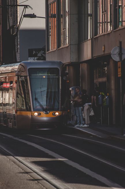 A modern, metallic silver tram with a sleek, curved front design is seen through the round mirror of a vehicle, reflecting city street lighting at night. The tram's windows are illuminated, revealing
